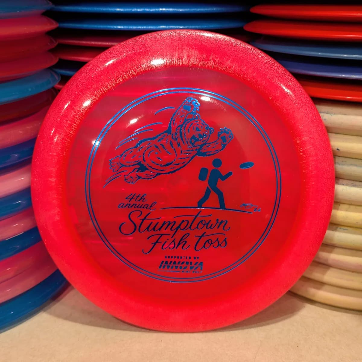 Blizzard Champion Boss, clear red, stamped with the 2026 Stumptown Fish Toss bear, leaning against the disc wall.
