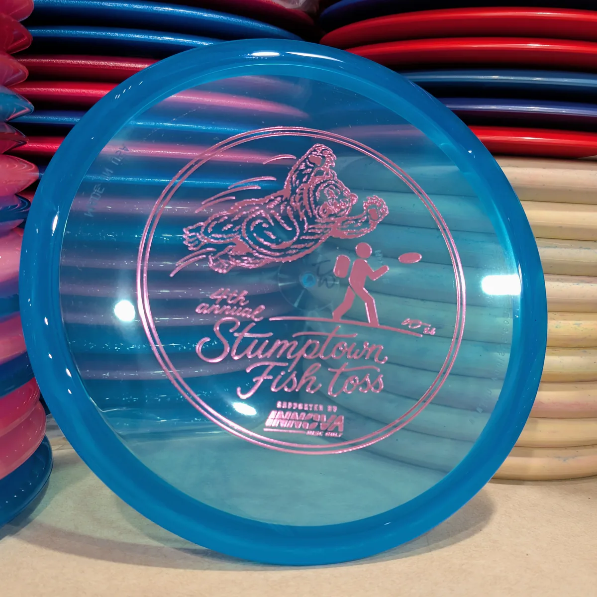 Champion Mako3, clear blue, stamped with the 2026 Stumptown Fish Toss bear, leaning against the disc wall.