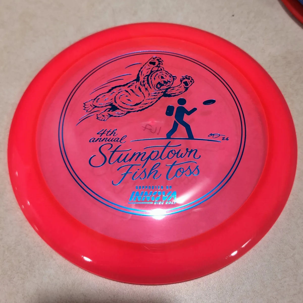 Champion Wraith, clear red, stamped with the 2026 Stumptown Fish Toss bear, leaning against the disc wall.