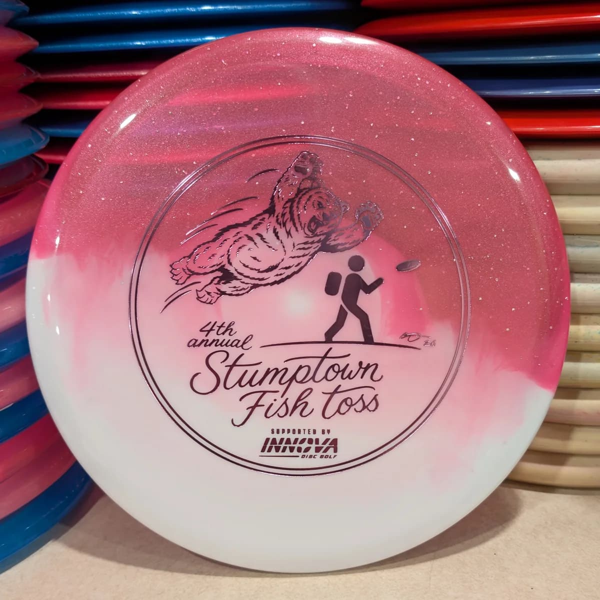Duo Roc3, pink and white, stamped with the 2026 Stumptown Fish Toss bear, leaning against the disc wall.