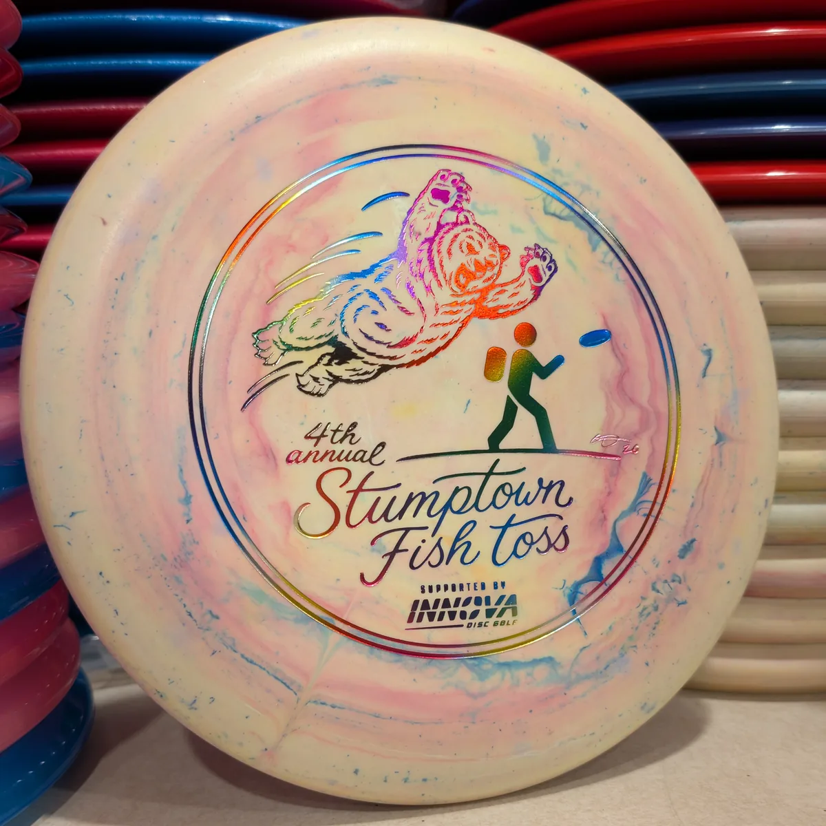 Galactic Pro Aviar, cream with a swirl of colour, stamped with the 2026 Stumptown Fish Toss bear, leaning against the disc wall.