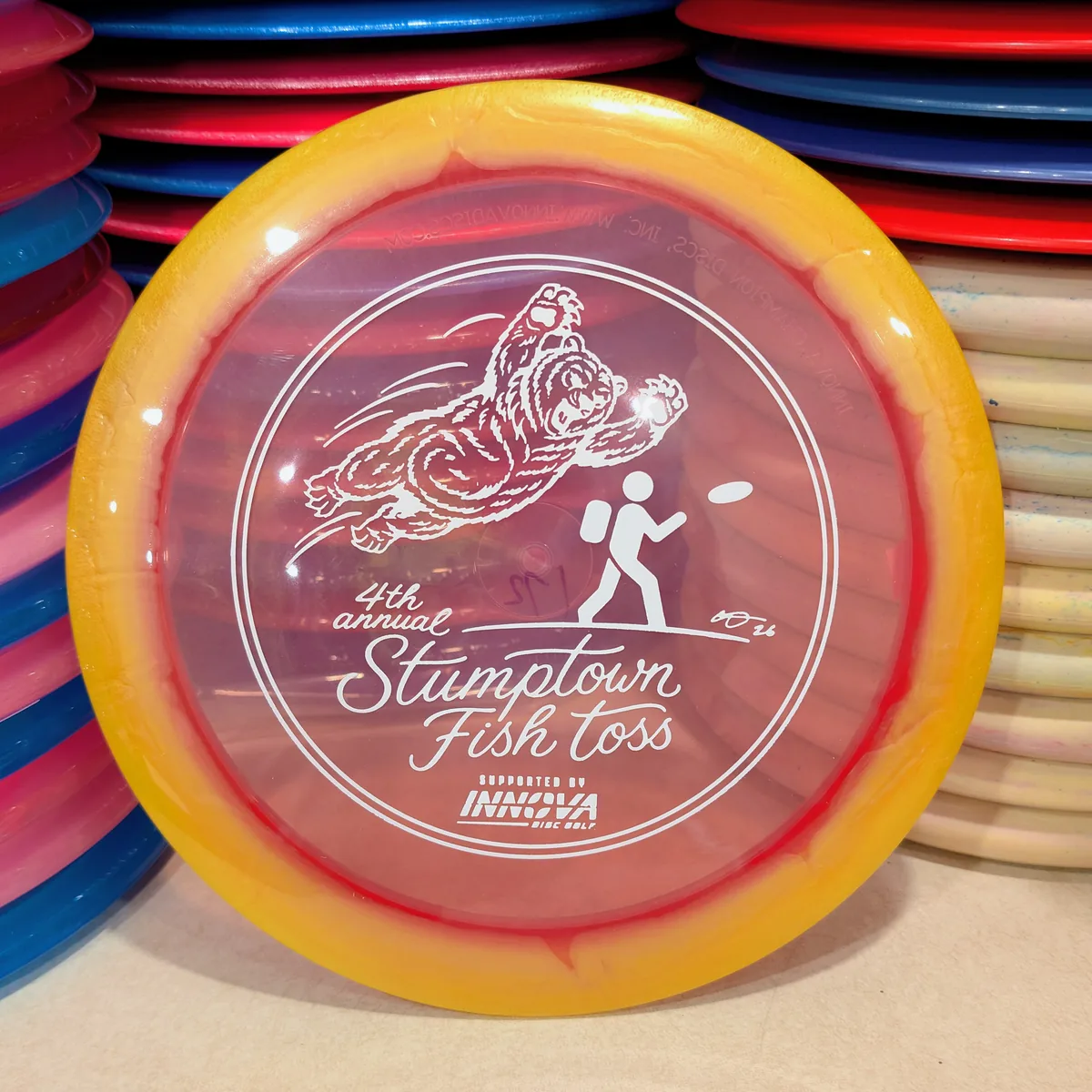 Halo Champion Roadrunner, clear with an orange rim, stamped with the 2026 Stumptown Fish Toss bear, leaning against the disc wall.