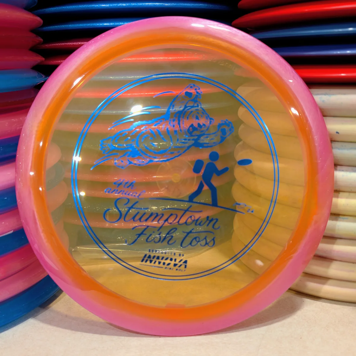 Halo Champion Wraith, orange with a pink rim, stamped with the 2026 Stumptown Fish Toss bear, leaning against the disc wall.