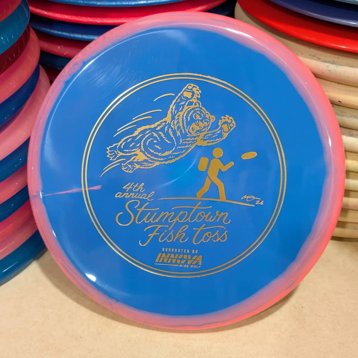 Halo Star Invader, blue with a pink rim, stamped with the 2026 Stumptown Fish Toss bear, leaning against the disc wall.