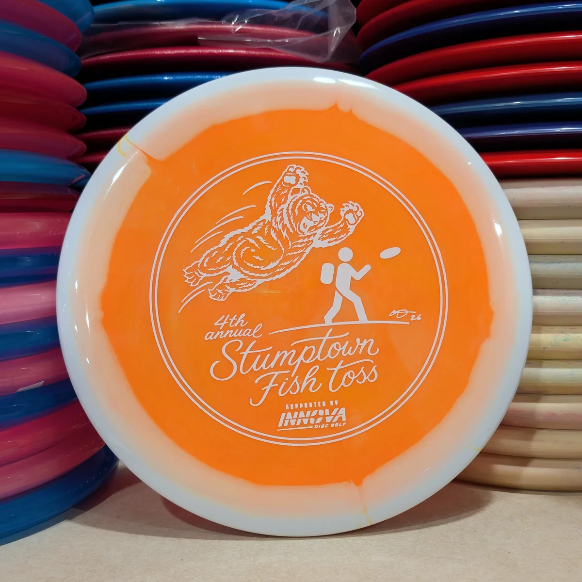 Halo Star Mako3, orange with a white rim, stamped with the 2026 Stumptown Fish Toss bear, leaning against the disc wall.
