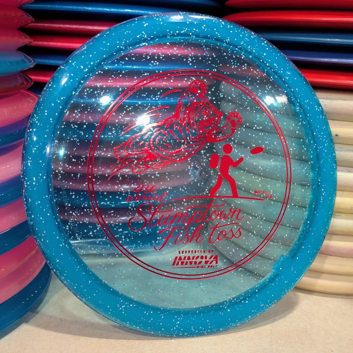Metalflake Champion TeeBird3, blue with metal flake sparkle, stamped with the 2026 Stumptown Fish Toss bear, leaning against the disc wall.