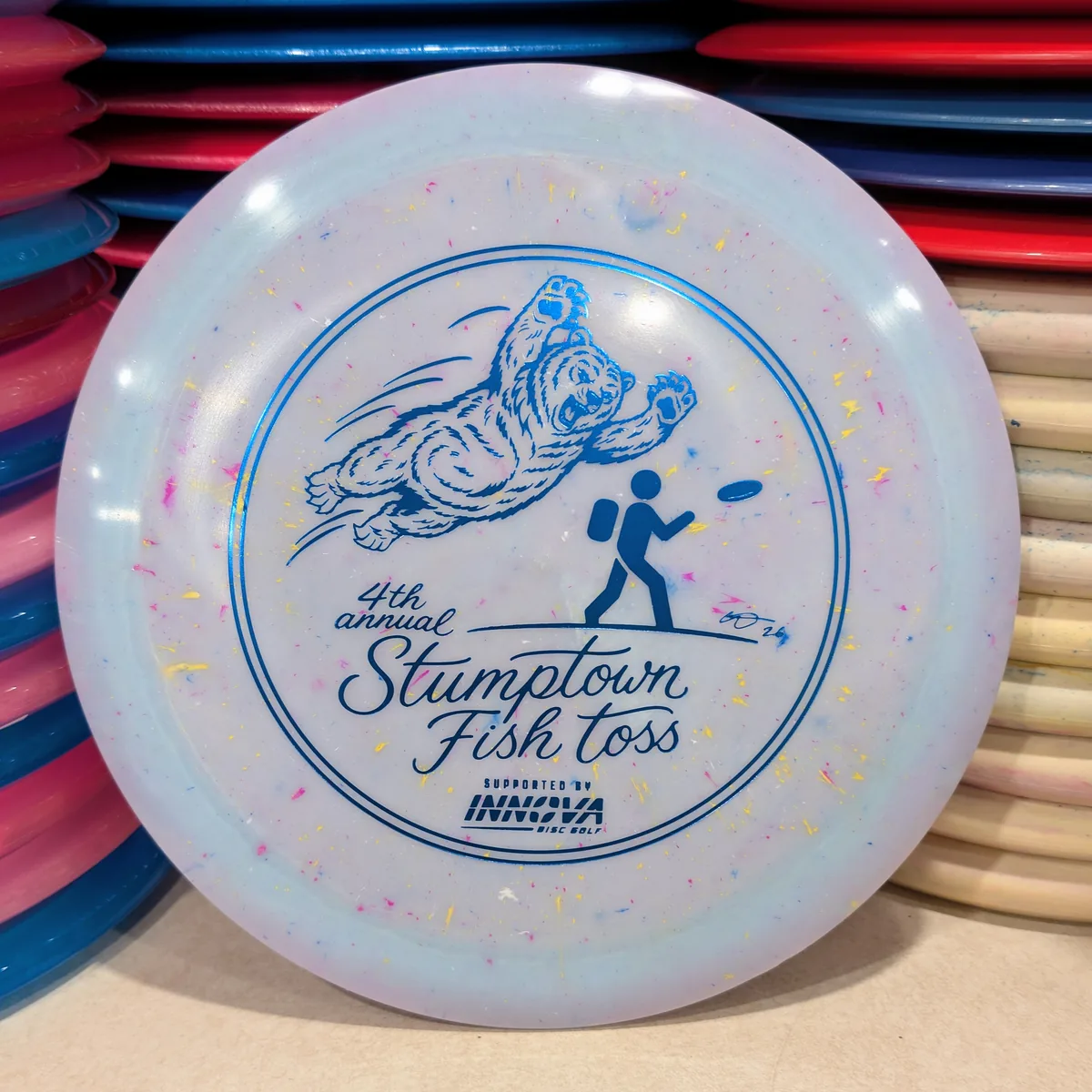 Splatter Star Destroyer, white with confetti flecks, stamped with the 2026 Stumptown Fish Toss bear, leaning against the disc wall.