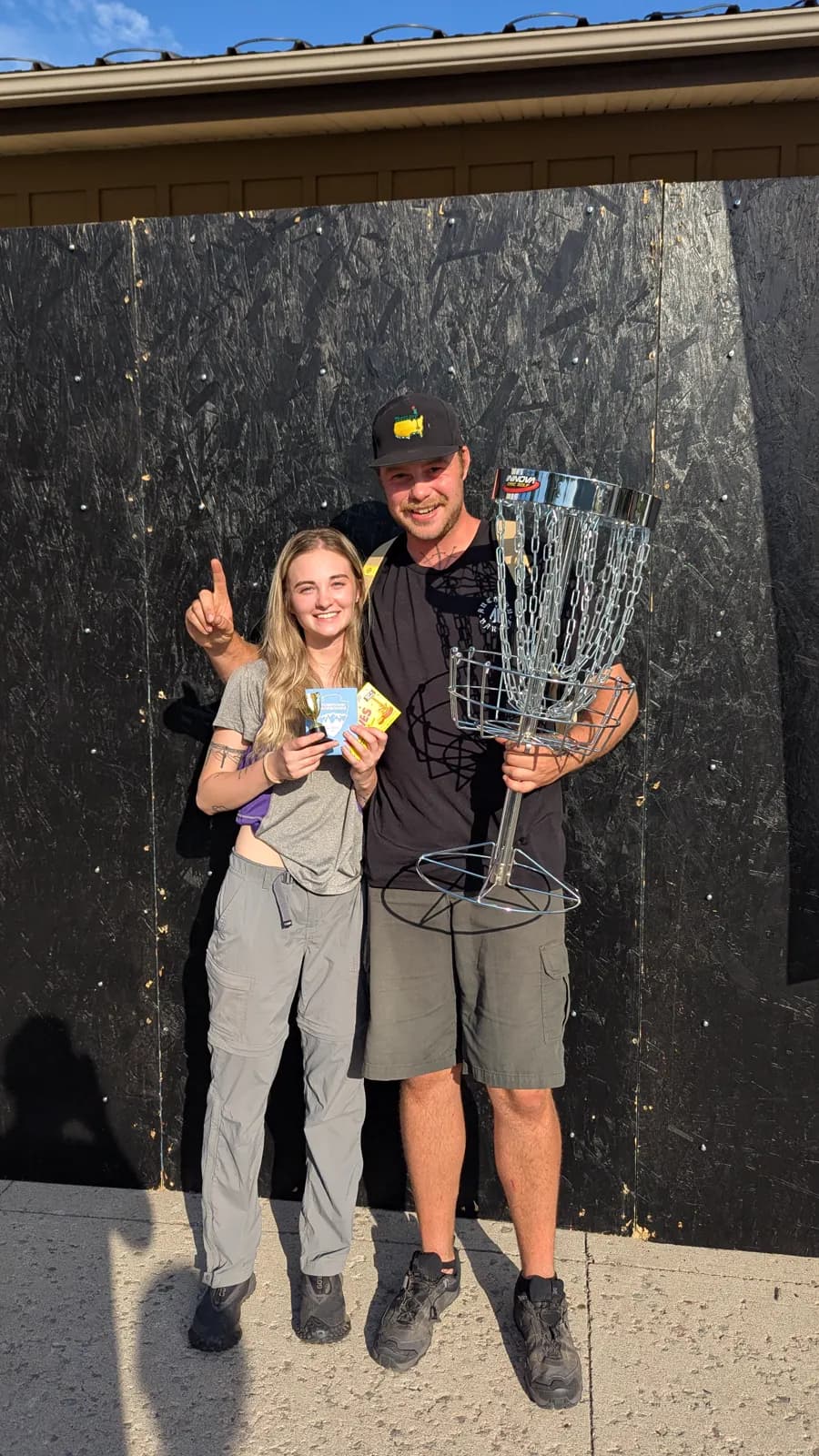 Two 2025 winners pose with a trophy and a disc golf basket.