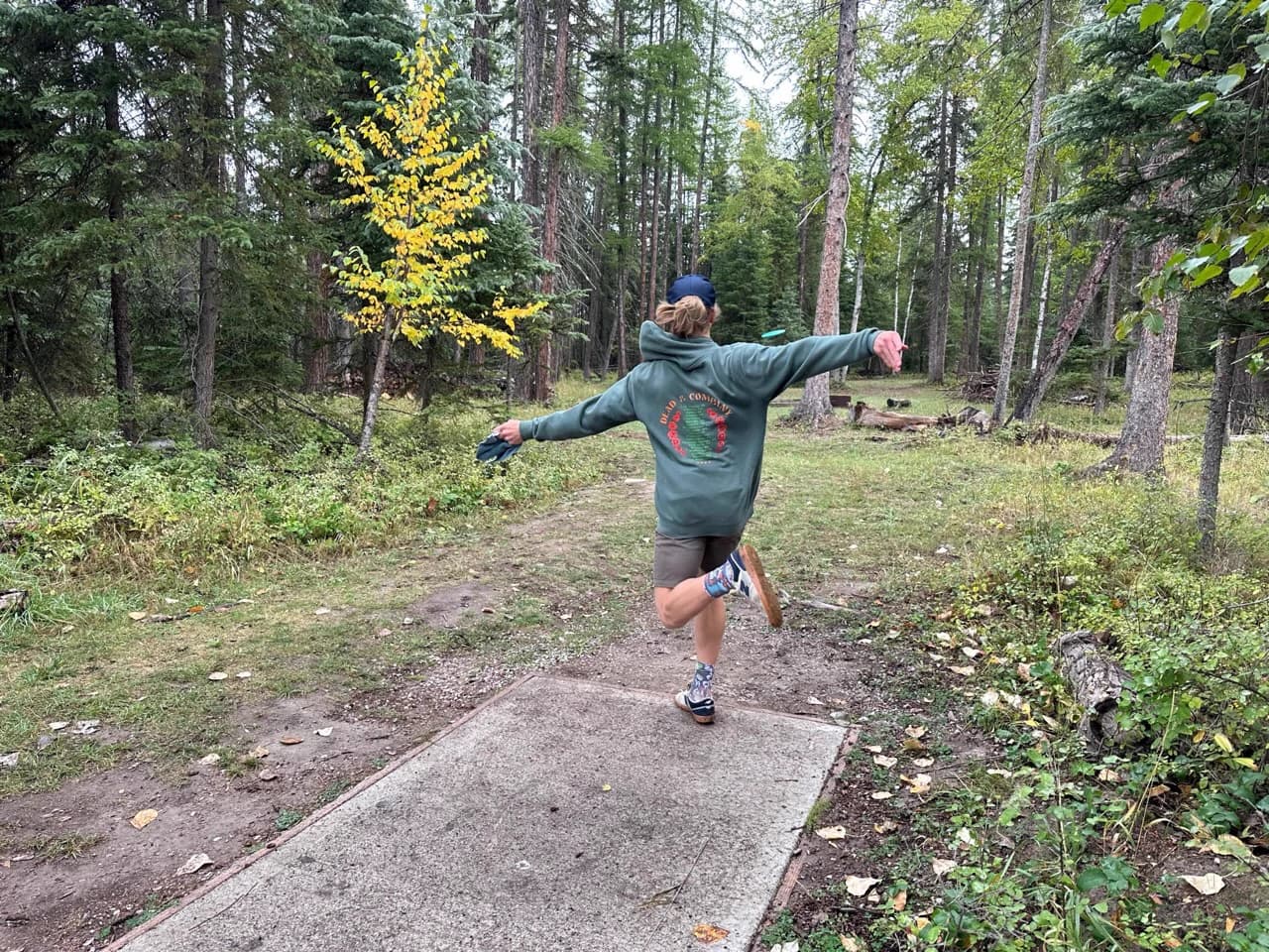 Seen from behind, a player balances on one leg after releasing a disc down a wooded fairway.