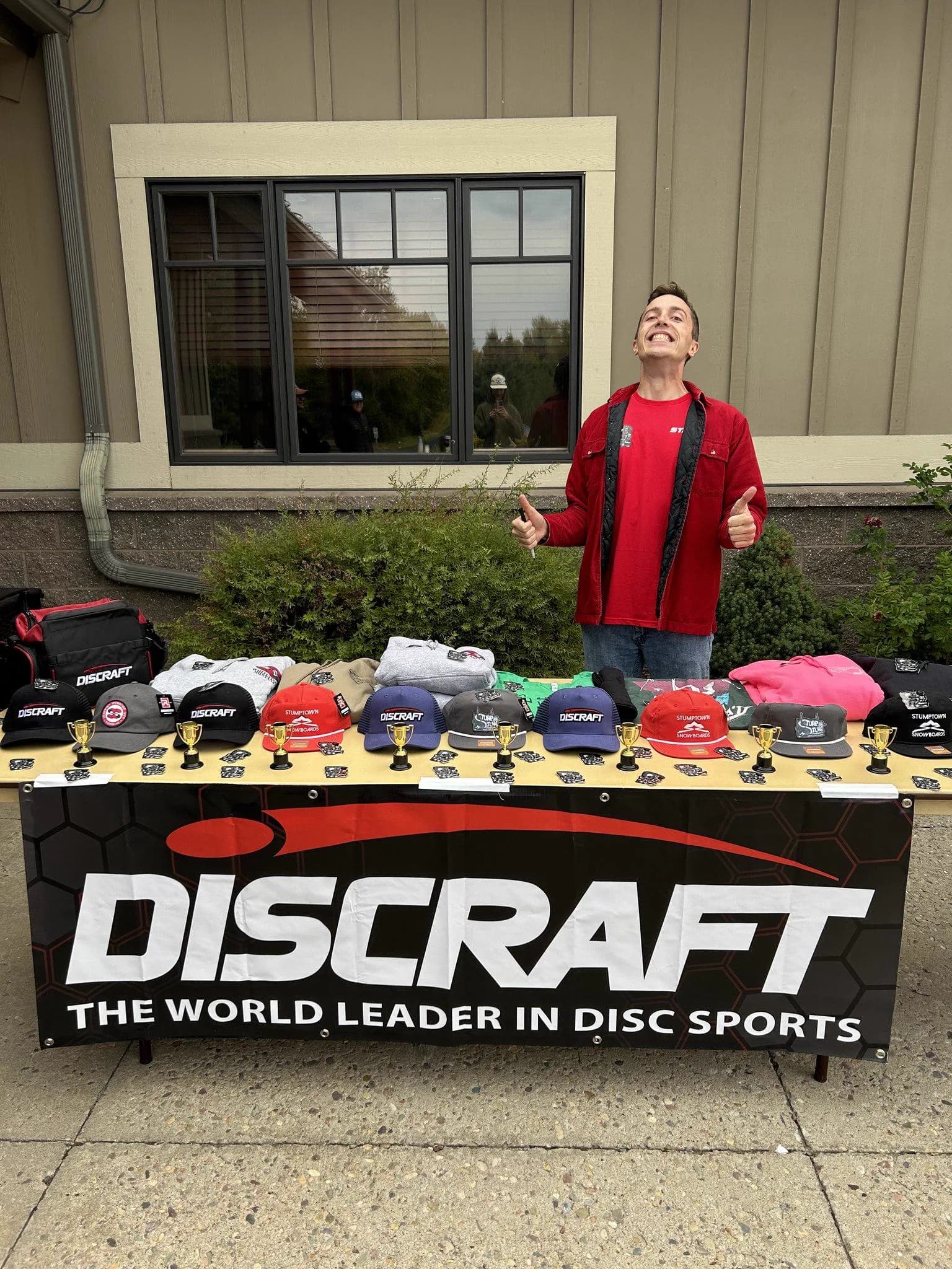 An organizer gives two thumbs up behind a prize table stacked with hats and prizes.