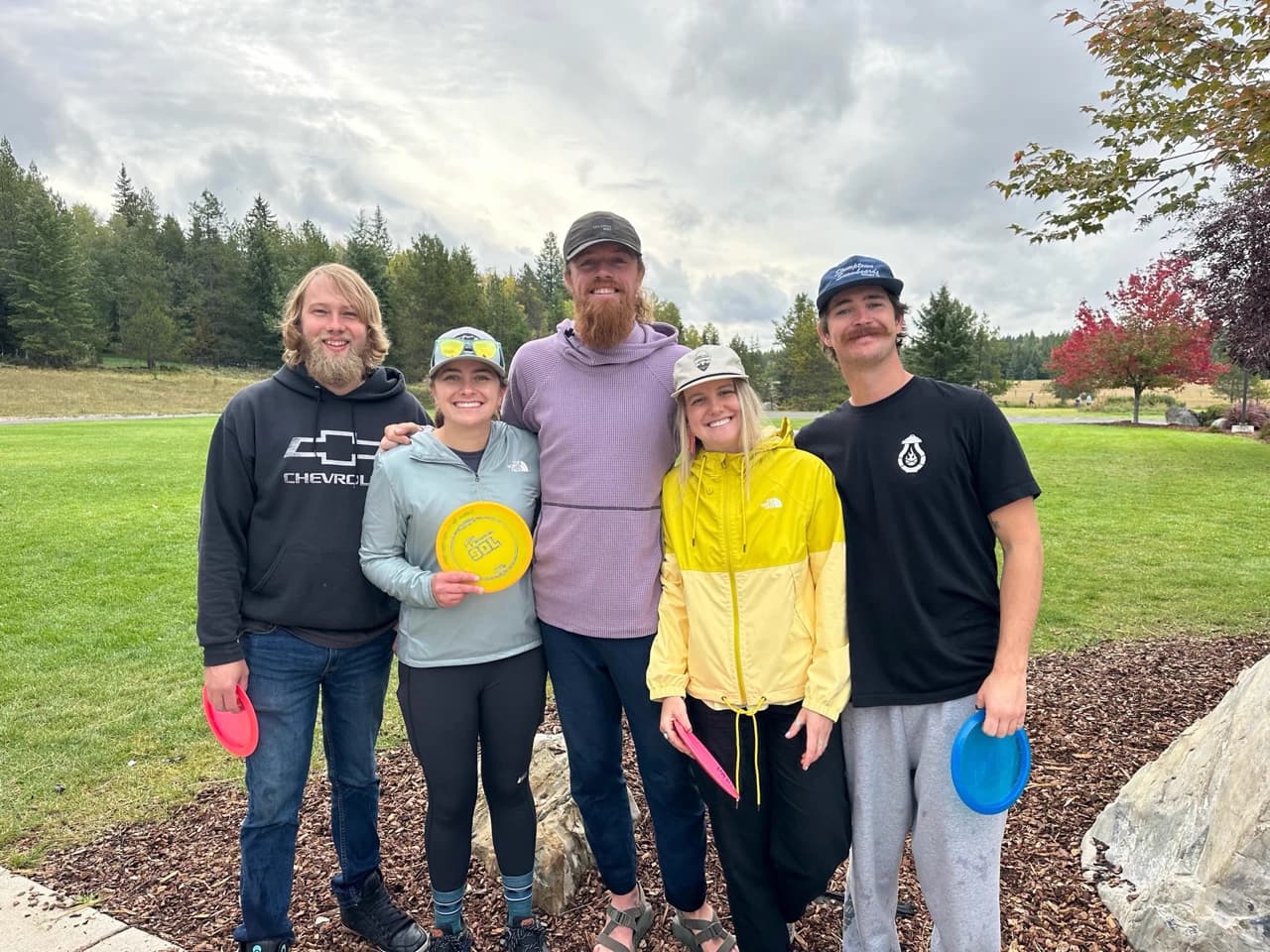 Five smiling players holding colorful discs pose on grass beneath a cloudy sky.