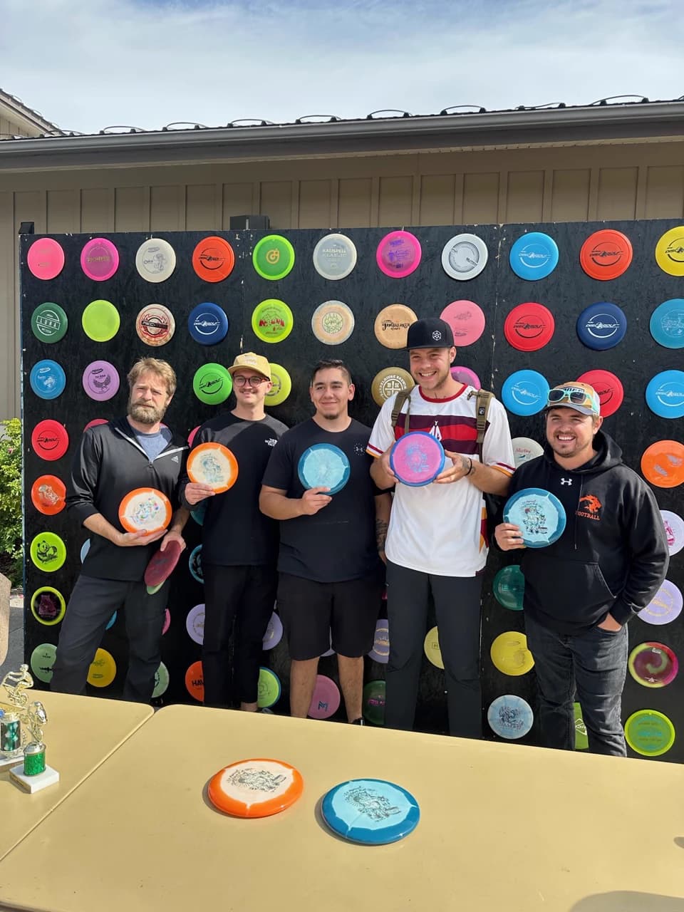 Five players hold stamped discs beside a trophy in front of the disc-covered wall.